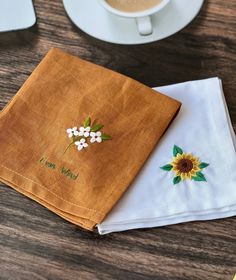 two napkins with embroidered flowers on them next to a cup of coffee and saucer