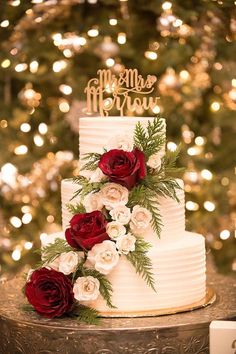 a white wedding cake with red and white flowers on top is sitting in front of a christmas tree