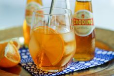 two glasses filled with liquid and orange slices on a plate next to bottles of soda