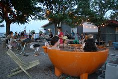 people are sitting in an orange bathtub on the beach at dusk with lights strung overhead