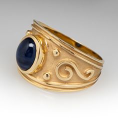 This artisanal ring is centered with one (1) oval cabochon cut natural sapphire set into a bezel setting. The ring features a high polished, raised scroll and beaded details atop a matted background. The ring measures 13.5mm at the top, rises 5.6mm above the finger, tapering to 3.7mm wide and 0.8mm thick at the base of the shank. It is currently a size 7. Timeless Cabochon Sapphire Ring, Formal 14k Gold Sapphire Ring With Oval Cabochon, Classic Oval Cabochon Sapphire Ring With Bezel Setting, Classic Sapphire Ring With Oval Cabochon Bezel Setting, Formal Sapphire Ring With Bezel Setting And Oval Cabochon, Yellow Gold Sapphire Ring With Bezel Setting, Oval Cabochon, Heirloom Style Sapphire Ring With Oval Cabochon, Heirloom Oval Cabochon Sapphire Ring, Heirloom Sapphire Ring With Oval Cabochon