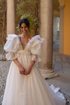 a woman in a white dress standing next to pillars and smiling at the camera with her hands on her hips