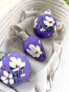 three purple ornaments with white flowers on them sitting in a wicker basket next to a towel