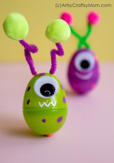 two toys with fake eyes are sitting on a pink surface and one is green, the other is purple