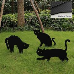 three black cats standing in the grass near some trees and bushes with a sign that says made of plastic