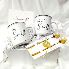 two coffee mugs in a gift box with some gold spoons on the side