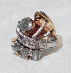 "This great, vintage two-tone ring shows detail and flair typical of the 1940s. The asymmetrical design is composed yellow gold, white gold, and four light blue topaz gemstones. The white gold \"ribbon\" is chased with tiny orange blossoms and decorated with milgrain. It separates the three smaller stones from the one larger stone. The fanned yellow gold section, which shares the side with the larger stone, has two slits and a little curl in the corner. The asymmetry even extends to the shank, w Vintage White Gold Multi-stone Diamond Ring, Vintage Multi-stone Diamond Ring In White Gold, Vintage White Gold Multi-stone Ring, Vintage Multi-stone Diamond Ring Collectible, Vintage Multi-stone Diamond Ring For Collectors, Vintage 14k Gold Rings With Accent Stones, Vintage Diamond Rings With Accent Stones, Vintage Topaz Ring With Diamond Accents For Anniversary, Vintage Diamond Topaz Ring With Accent Stones