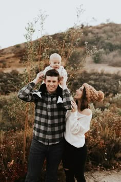 a man holding a baby while standing next to a woman in front of a tree