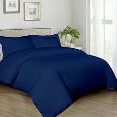 a bed with blue sheets and pillows in a white room next to a window on a hard wood floor