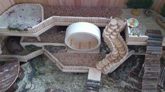 a toy house made out of wooden logs and sand in a garden area with plants