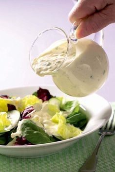 someone pouring dressing into a salad in a white bowl on a green tablecloth with silverware