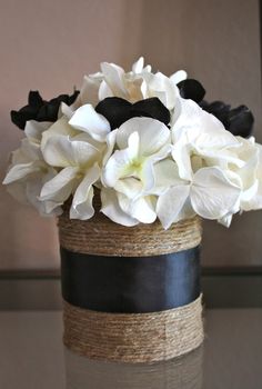 white flowers are in a black vase on a glass table top with rope wrapped around it