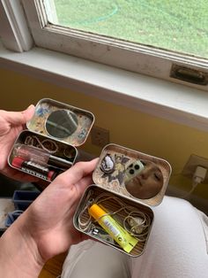 a person holding two small tins filled with various items in front of a window