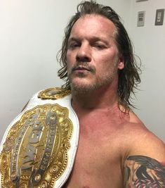 a man with long hair wearing a gold and white wrestling ring around his neck, standing in front of a wall