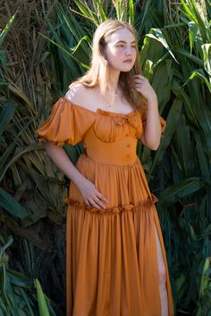 a woman in an orange dress standing next to some tall grass and plants with her hands on her hips