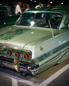 an old green car parked in a parking lot with other cars behind it at night