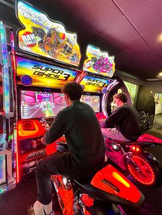 two men playing video games on motorcycles in front of neon signs and arcade machines at the same time