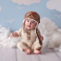 a baby wearing a pilot's hat and sitting on a cloud covered floor with clouds in the background