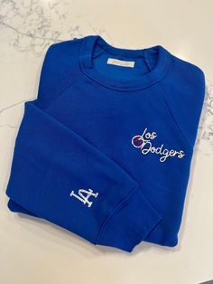 Whether you're a die-hard Dodgers fan or rooting for another team, this sweatshirt is the perfect addition to your game day wardrobe. Perfect for opening week festivities at the stadium or cozying up at home while cheering on your favorite team. This raglan crewneck sweatshirt is crafted from premium materials and embroidered with white, ecru and red thread.  Gear up for the season in style and comfort with a custom sweatshirt - the ultimate statement piece for any sports enthusiast. *Model is w Blue Sports Top With Logo, Sporty Blue Sweatshirt With Embroidered Logo, Blue Sports Sweatshirt With Embroidered Logo, Blue Varsity Sweatshirt With Embroidered Logo, Blue Sweatshirt With Embroidered Logo For Sports, Game Day Embroidered Crew Neck Sweatshirt, Collegiate Blue Top With Embroidered Logo, Crew Neck Sweatshirt With Embroidered Logo For Sports, Throwback Blue Sweatshirt For Game Day