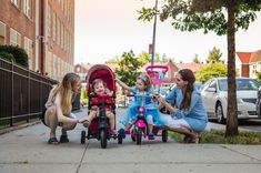 three women and one child are playing with their stroller on the sidewalk in front of some buildings