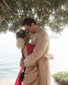 a bride and groom kissing under a tree by the water on their wedding day in india