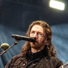 a man with long hair and beard holding a guitar in front of a microphone on stage