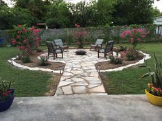 a stone patio with chairs and flowers in the back yard, next to a fire pit