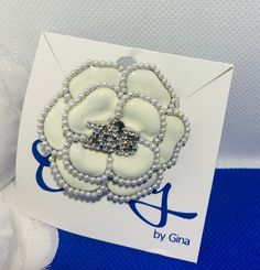 a white flower brooch with pearls on it's center and the words by gina written in blue