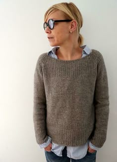 a woman standing in front of a white wall wearing a brown sweater and blue jeans