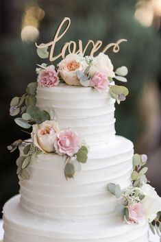 a white wedding cake with pink flowers and the word love on top is sitting on a table