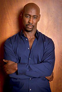 a man with his arms crossed standing in front of a wooden wall wearing a blue shirt