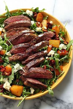 a yellow bowl filled with steak and salad on top of a marble countertop next to a fork