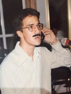 a man wearing glasses and a moustache talking on a cell phone with a microphone in front of him