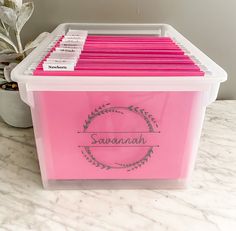 a pink storage box filled with files on top of a white countertop next to a potted plant