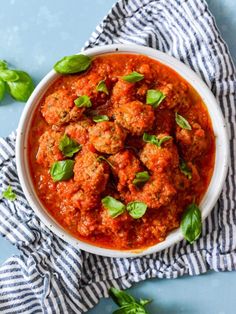 a white bowl filled with meatballs covered in marinara sauce and fresh basil leaves