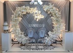 a white wedding arch with flowers and chairs on the floor in front of a backdrop