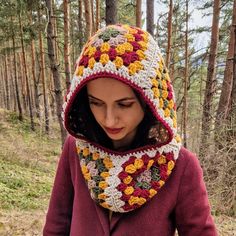 a woman wearing a crocheted hat and scarf in the woods with pine trees behind her