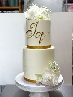 a three tiered cake with white flowers and gold lettering on the top is sitting on a pedestal