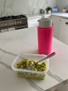 a plastic container filled with green pasta next to a pink water bottle on a counter