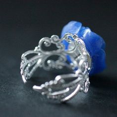 "A handmade cobalt blue resin rose has then been set upon an silver plated filigree adjustable ring base in your choice of finish in this romantic flower ring that is made to be adjustable from size 6 and up. Flower Size: 19mm To see this ring in all available colors, check here: http://etsy.me/1gr9dLc The base for this handmade ring is adjustable, which makes this delicate rose ring comfortable no matter the size of the wearer's fingers (size 6 and up) as well as ideal for switching from finger Blue Flower Ring For Gift, Unique Blue Flower Ring For Gift, Adjustable Blue Flower Ring, Elegant Adjustable Blue Flower Ring, Elegant Blue Adjustable Flower Ring, Adjustable Blue Flower Ring For Gifting, Adjustable Blue Flower Ring Gift, Handmade Blue Flower Ring, Resin Rose