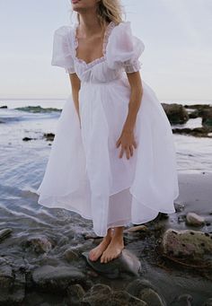 a woman in a white dress standing on rocks near the ocean with her hands behind her head