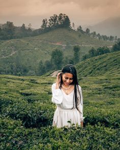 a woman standing in the middle of a lush green field with trees and hills behind her