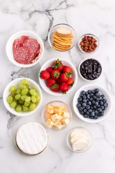 various fruits and cheeses are arranged in bowls on a marble countertop with crackers