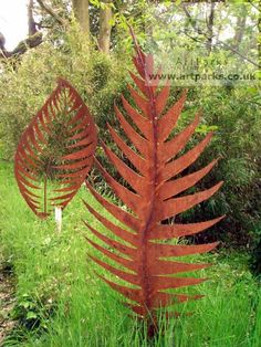 two large metal leaves in the grass near some trees and bushes, one is red