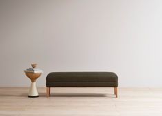 a small ottoman next to a wooden table with a vase on it and a white wall in the background