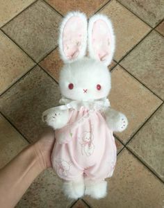 a hand holding a stuffed animal bunny in pink clothing on a tile floor next to a person's hand