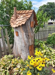 a tree stump with a house on it and some flowers in the foreground near a fence