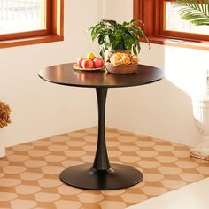 a small table with fruit on it in front of two windows and a potted plant