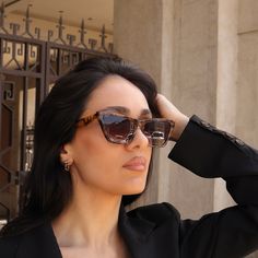 a woman wearing sunglasses and a black jacket is looking off into the distance with her hand on her head