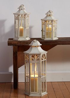 three white lanterns sitting on top of a wooden table next to a bench with candles in it